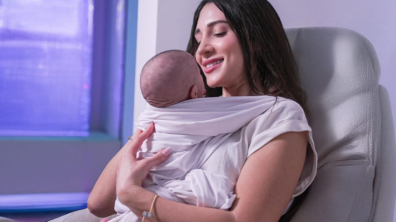 A smiling woman holds a baby close while seated in a cozy chair.