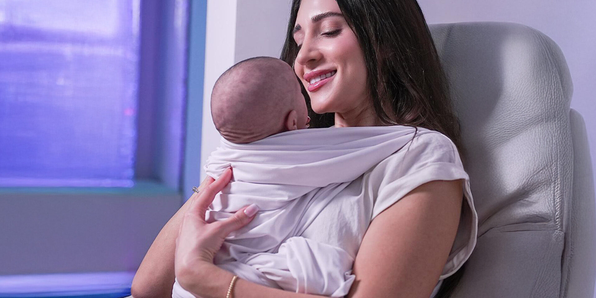 A smiling woman holds a baby close while seated in a cozy chair.
