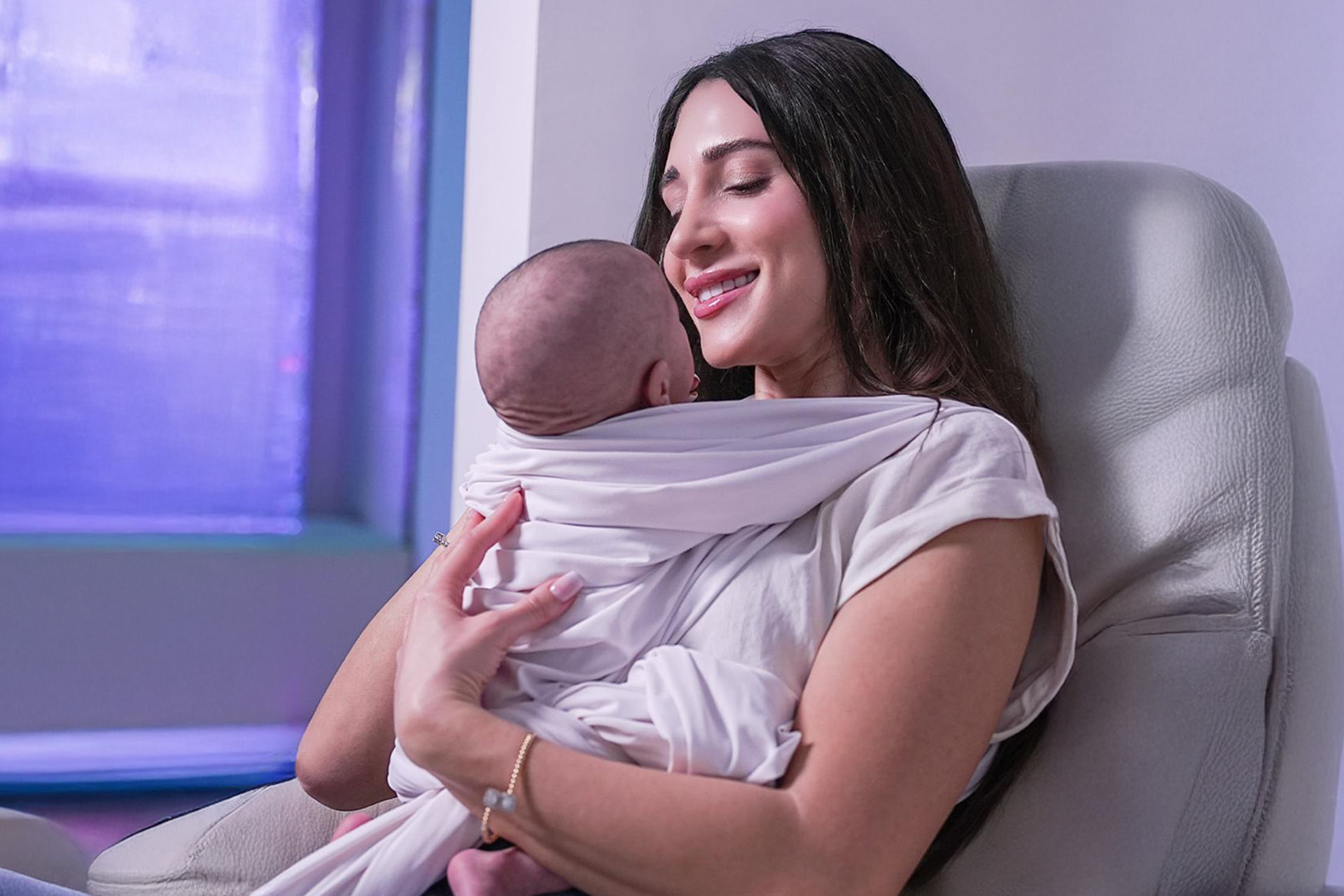 A smiling woman holds a baby close while seated in a cozy chair.